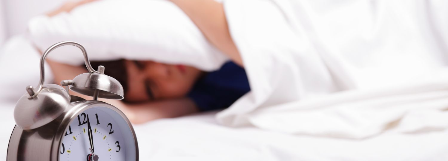 Man covering head with pillow next to alarm clock